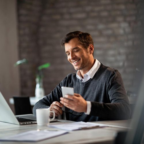 happy-businessman-texting-mobile-phone-while-working-laptop-office (2)